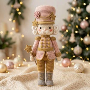 May include: A crocheted nutcracker doll in shades of soft pink and gold. The figure wears a tall hat with a rose and star accents, a jacket with gold buttons, and gold trousers. It holds a star-tipped wand and wears grey boots with laces.