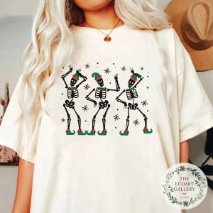 May include: White t-shirt featuring a design of three skeletons dressed as elves dancing. The skeletons are wearing green hats and red shoes. The background is white with snowflakes.