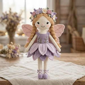 May include: A handcrafted crochet doll featuring light brown yarn hair styled in two braids, adorned with a floral crown. The doll wears a lavender dress with a leaf-shaped skirt, light pink fairy wings, and matching lavender ballet-style shoes.