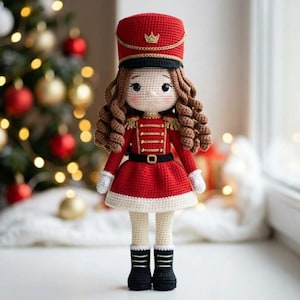 May include: A crocheted nutcracker doll featuring a red uniform with gold trim, a matching red hat with a gold crown emblem, and long brown curly hair. The doll wears a black belt, white gloves, cream-coloured leggings, and black boots with white laces.