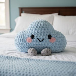 May include: A light blue cloud-shaped plush pillow made of chunky yarn. The pillow features two black plastic safety eyes, a stitched black smile, and two round pink cheeks. Two small, textured gray feet are attached to the bottom of the cloud.