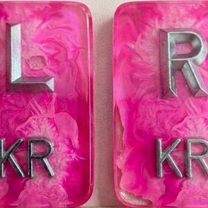May include: Two rectangular resin plaques featuring a vibrant pink and white marbled pattern. The left plaque displays a large silver letter L above the letters KR. The right plaque displays a large silver letter R above the letters KR.