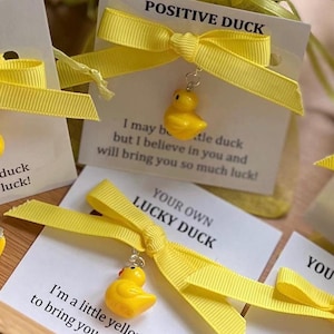 Puede incluir: Un conjunto de cuatro patitos de goma amarillos con una cinta amarilla atada alrededor de cada uno. Cada encanto tiene una pequeña etiqueta adjunta con un mensaje sobre la buena suerte. Las etiquetas dicen "Pato Positivo", "Tu Propio Pato de la Suerte", y "Soy un pequeño pato amarillo para traerte mucha suerte."