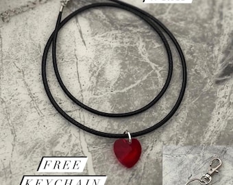 Könnte beinhalten: Eine schwarze Choker-Kette mit einem roten Herz-Anhänger. Die Kette liegt auf einer grauen, strukturierten Oberfläche. "FREE GIFT WRAP" und "FREE KEYCHAIN" sind in weißer Schrift auf schwarzen Rechtecken geschrieben.