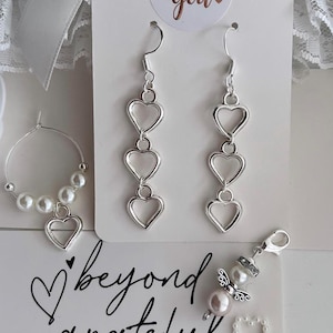 Può includere: Un set di orecchini a forma di cuore argentati, un braccialetto con ciondolo a forma di angelo di perla e un biglietto bianco con il testo "beyond grateful" e "thank you so much".