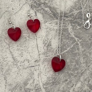 Puede incluir: Un collar de cadena de plata con un colgante de corazón de cristal rojo. También se incluyen pendientes de corazón de cristal rojo a juego.