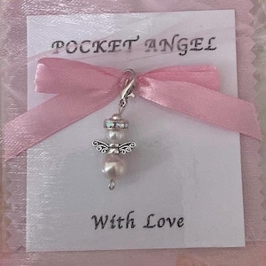 Puede incluir: Un amuleto de ángel blanco con halo y alas plateadas, colgando de una cadena plateada. El amuleto está en una tarjeta blanca con cinta rosa y el texto "Pocket Angel" y "With Love".