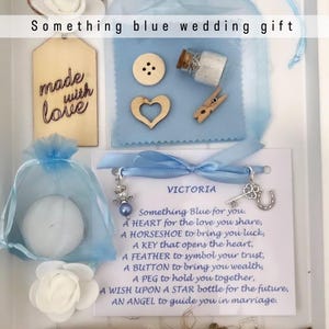 Puede incluir: Caja de regalo de boda 'Something Blue'. Contiene varios objetos pequeños, como una etiqueta de madera con 'made with love', un corazón, un botón, una botella pequeña, una pinza, una pluma y una tarjeta con un poema. Decorada con detalles azules.
