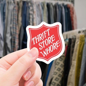 May include: A red and white sticker with the text "Thrift Store Whore" in a white font. The sticker is shaped like a shield.