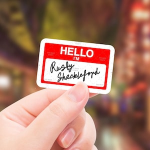 May include: A red and white name tag with the text "HELLO I'M Rusty Shackleford" written in black marker.