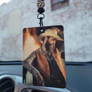 Puede incluir: Ambientador rectangular para coche con un retrato de un hombre con sombrero y chaleco de vaquero. El ambientador está suspendido de una cadena de cuentas con cuentas a cuadros en blanco y negro y un cierre negro. El fondo es un edificio de ladrillo borroso.