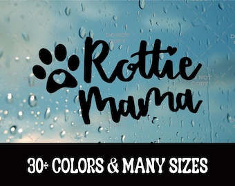 May include: A black vinyl decal featuring a paw print with a small heart cutout and the text "Rottie Mama" in a cursive script font. Below the graphic, a black banner displays the white text "30+ COLORS & MANY SIZES" in a bold, sans-serif typeface.
