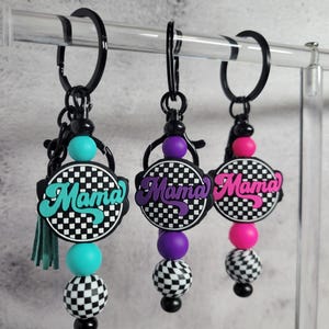 Puede incluir: Tres llaveros con la palabra "Mama" en verde azulado, morado y rosa, cada uno sobre un fondo a cuadros blanco y negro. Los llaveros tienen herrajes negros, cuentas de colores y una borla.