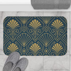 Peut inclure: Un tapis de bain rectangulaire avec un motif art déco répétitif. Le motif présente des formes dorées en forme d'éventail et des éléments géométriques sur un fond bleu canard foncé. Des pantoufles grises sont dans le coin inférieur gauche.