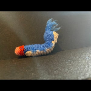 May include: A blue and gray crocheted plush toy shaped like a worm with a red and pink crocheted head.