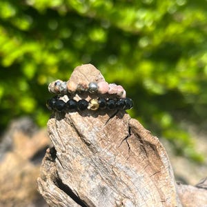 Puede incluir: Tres anillos de cuentas apilados. El anillo superior tiene cuentas grises, rosas y plateadas. El anillo del medio es negro con una cuenta dorada. Los anillos se muestran sobre un trozo de madera desgastada, con un fondo verde borroso.