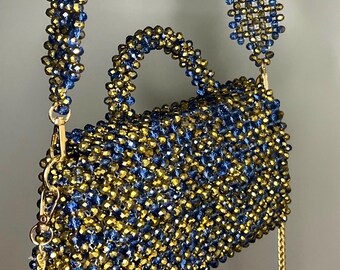 May include: A beaded handbag with a rectangular shape, featuring a handle and a shoulder strap. The bag is adorned with blue and gold beads, creating a textured surface. A gold chain is attached to the bag for additional carrying options.