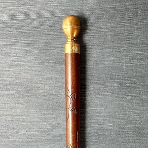 May include: A brown wooden walking cane with a gold-colored handle and tip. The cane features carved floral designs along its length. The top of the cane has a rounded gold-colored knob.