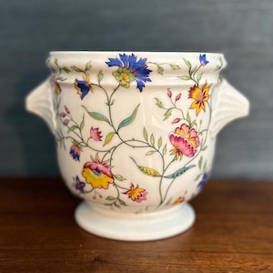 May include: White ceramic planter with floral pattern in shades of blue, yellow, pink, and green. The planter has two small handles on the sides and a wide base.