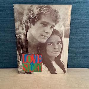 以下が含まれることがあります： 男性と女性の白黒写真が特徴のヴィンテージ本の表紙。「LOVE STORY」のタイトルが大きくカラフルな文字で印刷されています。表紙は青色の背景に配置されています。