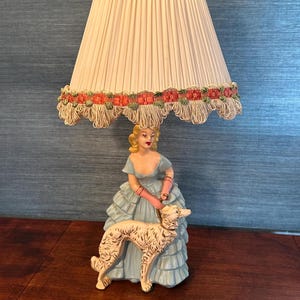 May include: A white lampshade with a pink and green trim sits atop a ceramic lamp base. The base features a woman in a blue dress holding a white dog.