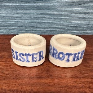 May include: Two small, off-white ceramic ring holders with blue lettering. One reads "SISTER" and the other reads "BROTHER". The ring holders are on a dark wooden surface, with a blue textured background.