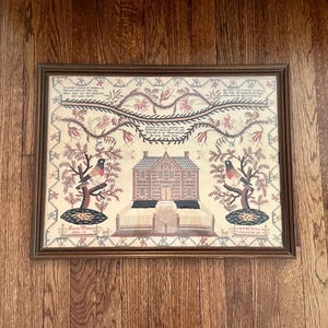 May include: Framed antique needlepoint sampler featuring a central house flanked by trees with birds. The sampler has a brown wooden frame and is set against a wood grain background. The needlepoint uses a variety of colors and patterns.