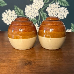 Puede incluir: Un par de saleros y pimenteros de cerámica. Cada salero y pimentero tiene un diseño redondeado en forma de barril con una base beige claro, una banda marrón medio y una parte superior marrón más oscura. Los saleros y pimenteros tienen un acabado brillante.