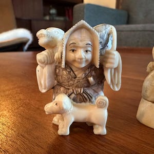 May include: A small off-white carved figurine depicting a person wearing a patterned garment and a pointed hat, carrying a bundle on their shoulders with a small dog standing at their feet, positioned next to a larger partial carved figure.