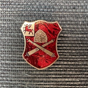 May include: A vintage shield-shaped pin with a red background and silver trim. The pin features a winged horse, a shield, and crossed cannons. The pin is likely made of metal and has a textured appearance.