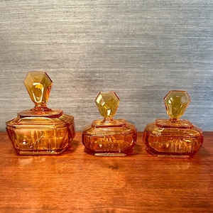 May include: Three amber-colored glass jars with faceted sides and geometric, gem-shaped lids. The jars are arranged in a row, decreasing in size from left to right. The jars sit on a wooden surface.