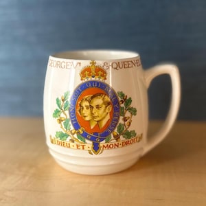 Może przedstawiać: Kremowy ceramiczny kubek z uchwytem, ​​z królewskim herbem z portretami, koroną i tekstem. Kubek ma zaokrąglony kształt i białe wnętrze. Projekt zawiera czerwone, niebieskie i złote akcenty.