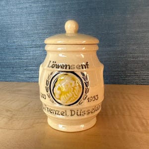 Peut inclure: Un pot en céramique beige avec un couvercle, portant l'inscription "Löwensenf" et un emblème circulaire avec une tête de lion. Le pot porte également les années "1903" et "1953" et le texte "Frenzel, Düsseldorf".