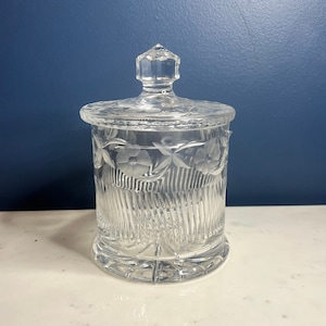 Könnte beinhalten: Eine klare Glasdose mit Deckel, verziert mit einem geätzten Blumendesign und einem strukturierten, gerippten Muster. Die Dose hat einen runden Boden und einen leicht konischen Körper.