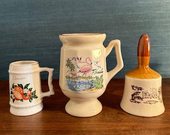 May include: Three ceramic Florida souvenir mugs. One mug features a pink flamingo, another has oranges, and the third is a bell with a dolphin and alligator design. All mugs have the word "Florida" on them.
