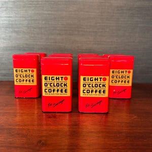 May include: Several red Eight O'Clock Coffee tins are arranged on a wooden surface. Each tin has a yellow label with black text that reads "Eight O'Clock Coffee" and "Freshly Roasted Ground to Order Coffee". The words "For Savings" are written in red.