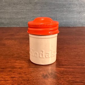 Può includere: Un piccolo contenitore per pellicola Kodak vintage. Il contenitore cilindrico ha un corpo bianco e un coperchio arancione brillante. La parola "Kodak" è in rilievo sul lato. Il coperchio è a vite. Un classico accessorio fotografico.