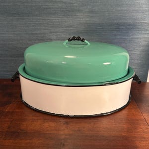 May include: A vintage enamelware baking dish with a teal lid and white base. The dish has a black rim and black handles.