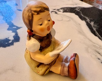 May include: A ceramic figurine of a young girl singing from a sheet of music. The girl is wearing a yellow dress, brown vest, and red hair ribbons. Her skin is fair, and her expression is serene. The figurine is a collectible item.