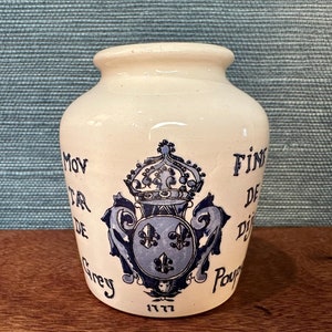 Könnte beinhalten: Eine weiße Keramikdose mit blauer und schwarzer Beschriftung und einem blauen und weißen Wappen. Die Dose trägt die Aufschrift "Fin de Dijon" und "Moutarde de Grey" mit dem Jahr 1777 am Boden.