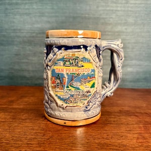 Könnte beinhalten: Eine Keramiktasse mit detailliertem Reliefdesign und einem griff in Form eines Astes. Die Tasse zeigt eine farbenfrohe Grafik von San Francisco, Kalifornien, mit Wahrzeichen. Der Rand und der Boden sind orange.