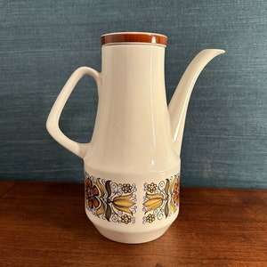 Puede incluir: Una cafetera de cerámica blanca con tapa marrón y un diseño floral en tonos naranja, amarillo y verde.
