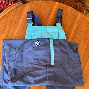 May include: Dark blue overalls featuring a light blue chest panel and shoulder straps. A circular BMW logo patch is centered on the chest. The garment includes a front pocket, two side pockets, and adjustable metal buckles on the shoulder straps.