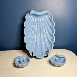 May include: A set of three light blue ceramic seashell-shaped dishes. The largest dish is a serving dish with a raised handle. The two smaller dishes are round and have a small indentation in the center.