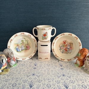 May include: A collection of Bunnykins children's dishware and figurines. The set includes two bowls, a handled cup, and a Bunnykins bank. The bowls and cup feature colorful illustrations of rabbits. The figurines include a squirrel, owl, and a rabbit.