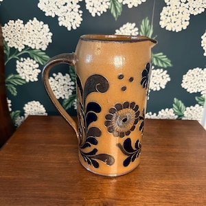 May include: A tan ceramic jug with a dark brown handle and rim. The jug is decorated with dark blue floral designs. The jug is sitting on a wooden surface.