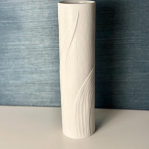 May include: A tall, cylindrical white ceramic vase with a textured surface and a subtle leaf design. The vase is approximately 10 inches tall and has a smooth, rounded top. The background is a muted blue.