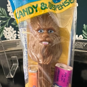 Könnte beinhalten: Ein brauner Plastik-Chewbacca-Charakter aus Star Wars ist an einem Süßigkeitenspender befestigt. Der Spender ist in Klarsichtfolie verpackt und enthält einen rosa-violetten Bonbonstick. Die Verpackung trägt die Aufschrift "Candy & Dispenser" und "Grape Candy" mit einem Nettogewicht von 29 oz (8,5 g).