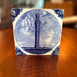 May include: A square ceramic tile featuring a blue and white transferware design. The center depicts the Pilgrim Memorial Monument in Provincetown, Mass. The corners are decorated with floral patterns, and the text reads Pilgrim Memorial Monument Provincetown Mass.