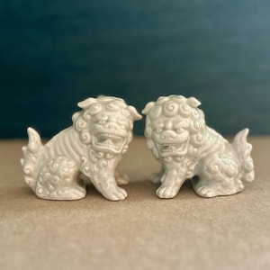 May include: Two off-white ceramic guardian lion figurines. Each lion is in a seated position with a detailed mane, open mouth, and a curled tail. The background is a muted teal color.